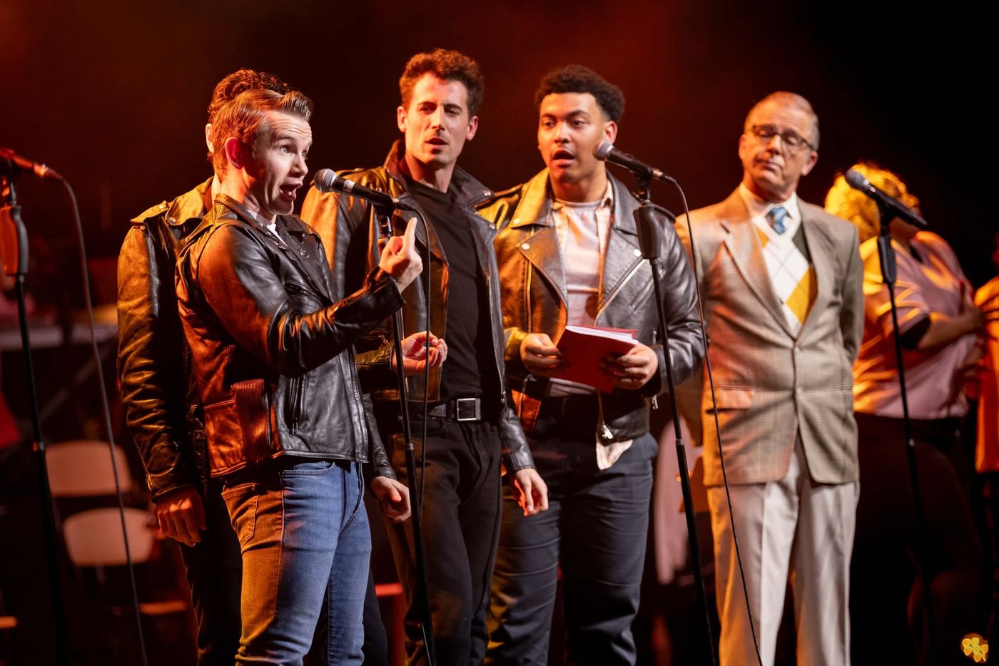 Four performers at microphones, one in a tan suit