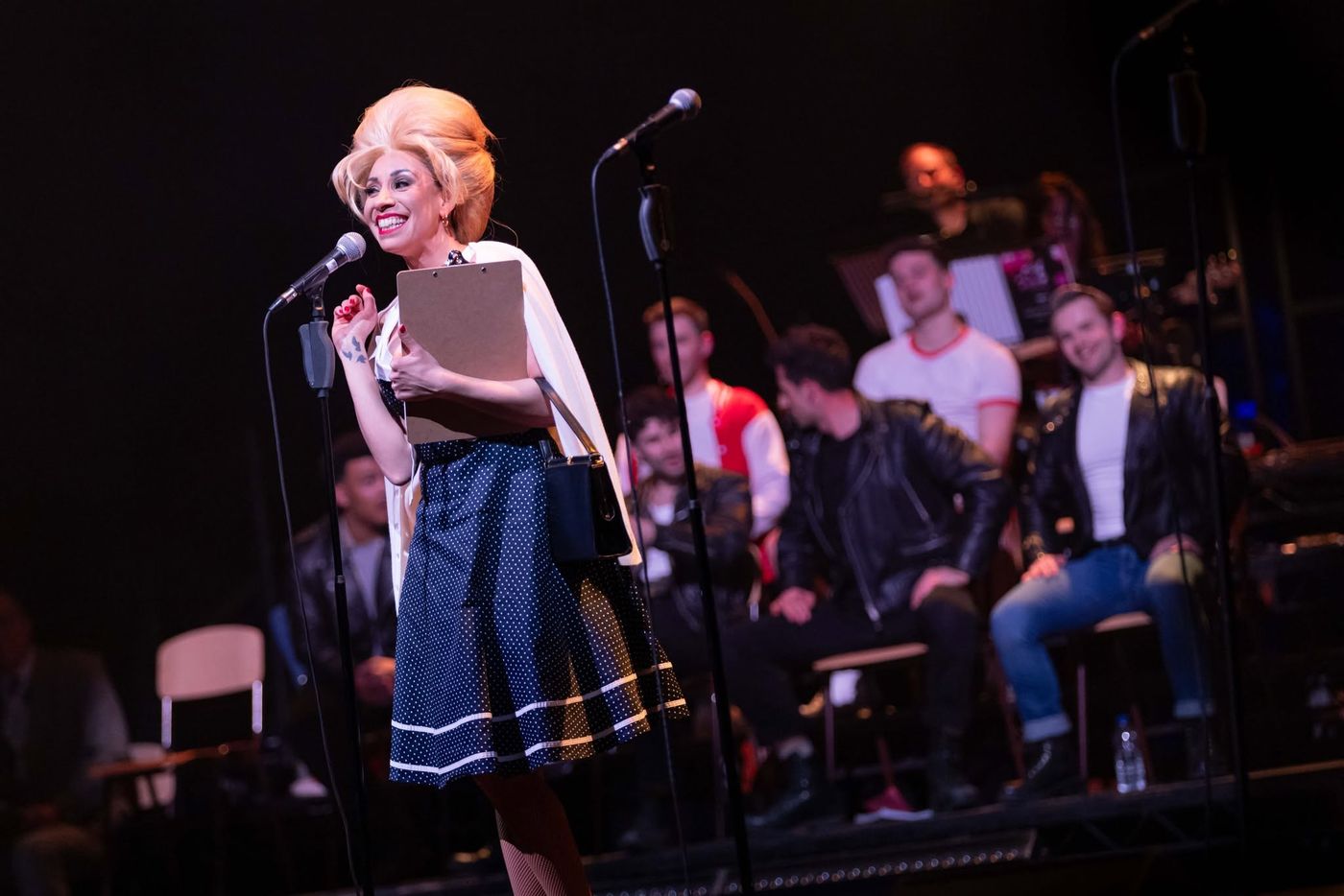 A performer singing downstage with the company seated behind