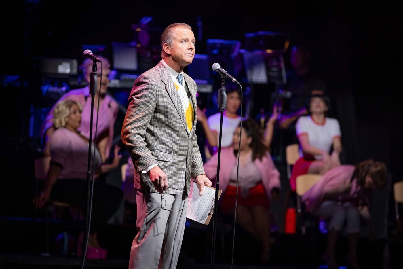 A performer in a grey suit at the microphone with the cast behind